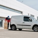 A delivery driver wearing a face mask unloads packages from a van outside a modern building.